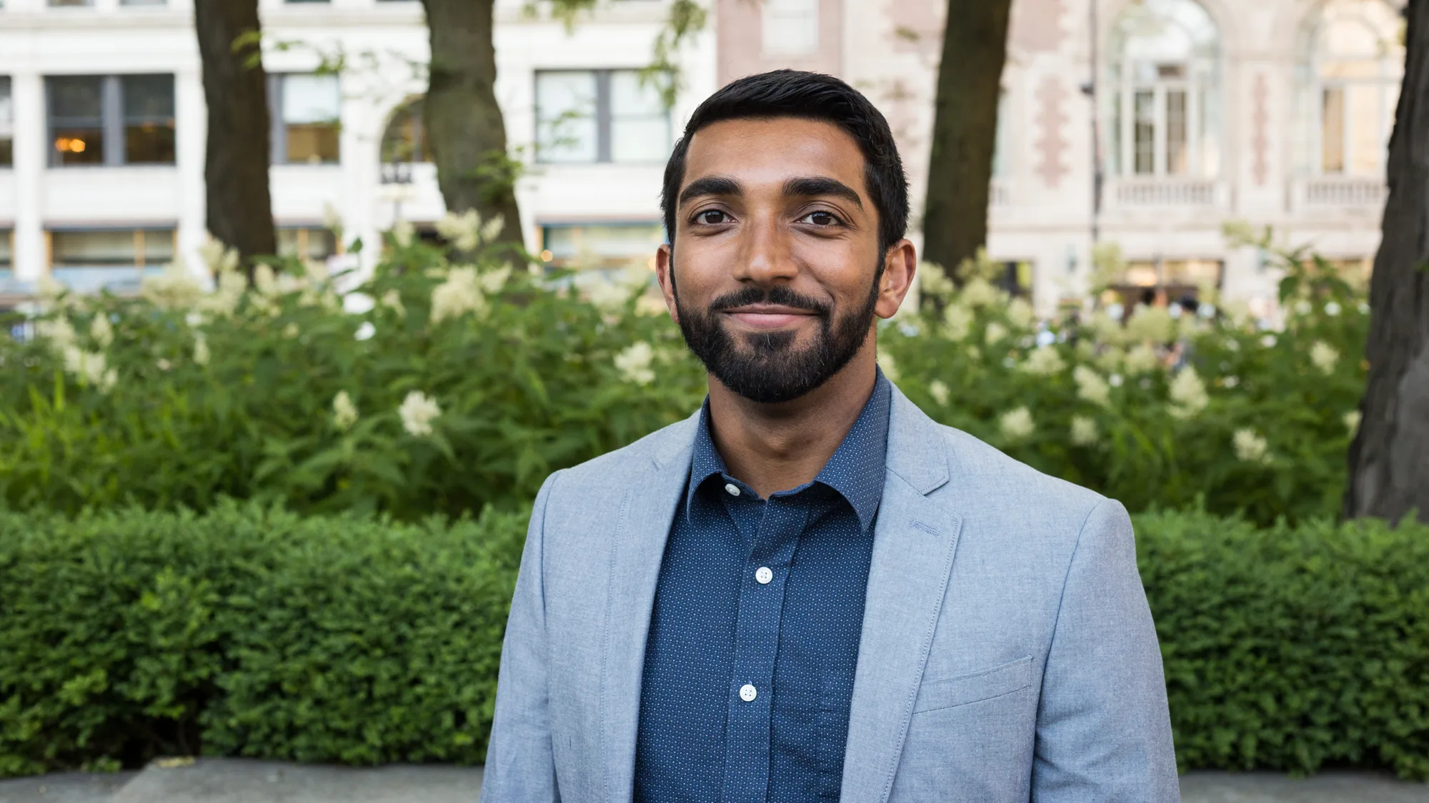 Headshot of Bejoy, wearing business casual attire with trees and foliage in the background.